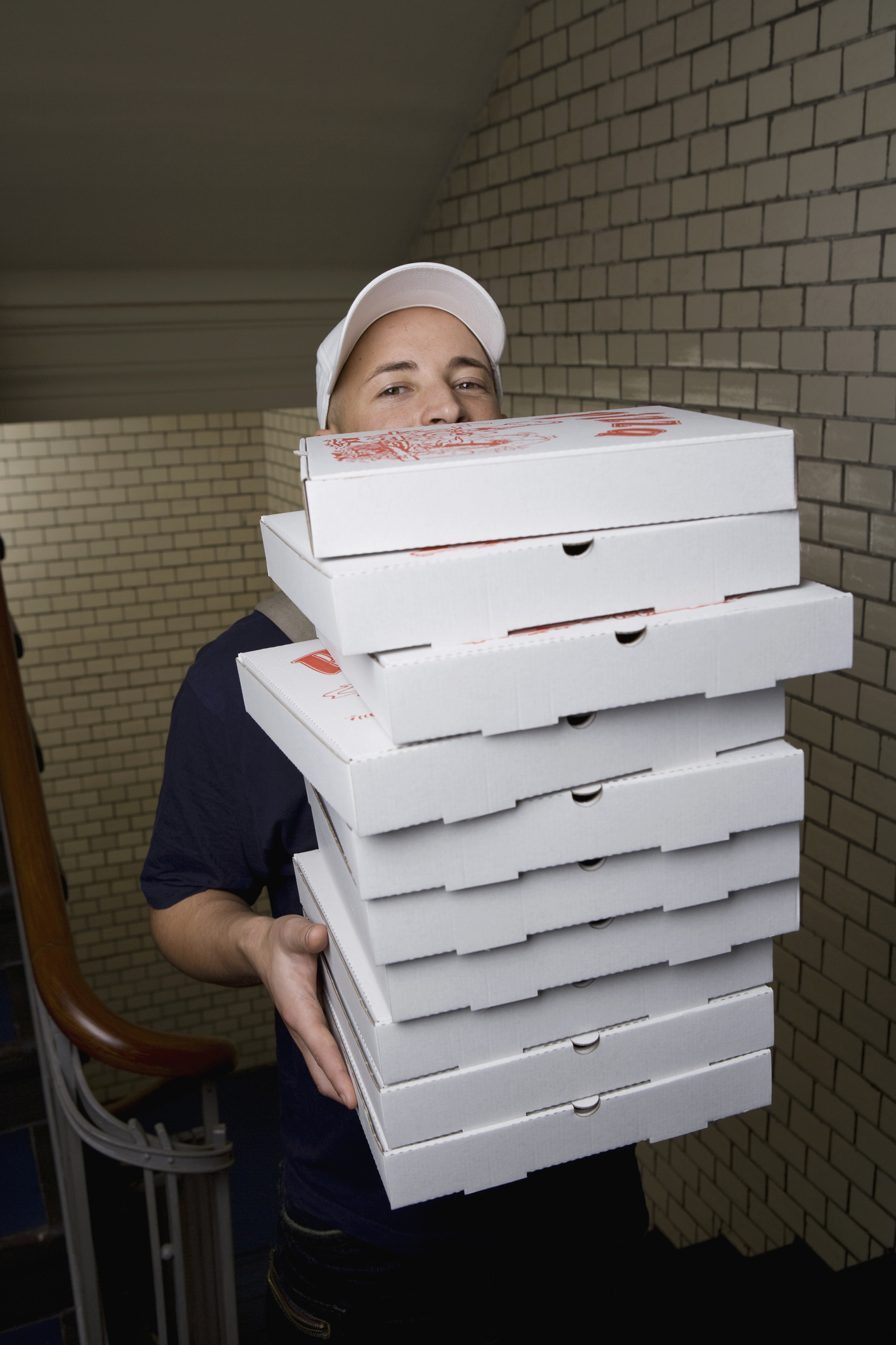 Person in a white cap carrying a large stack of pizza boxes up a staircase