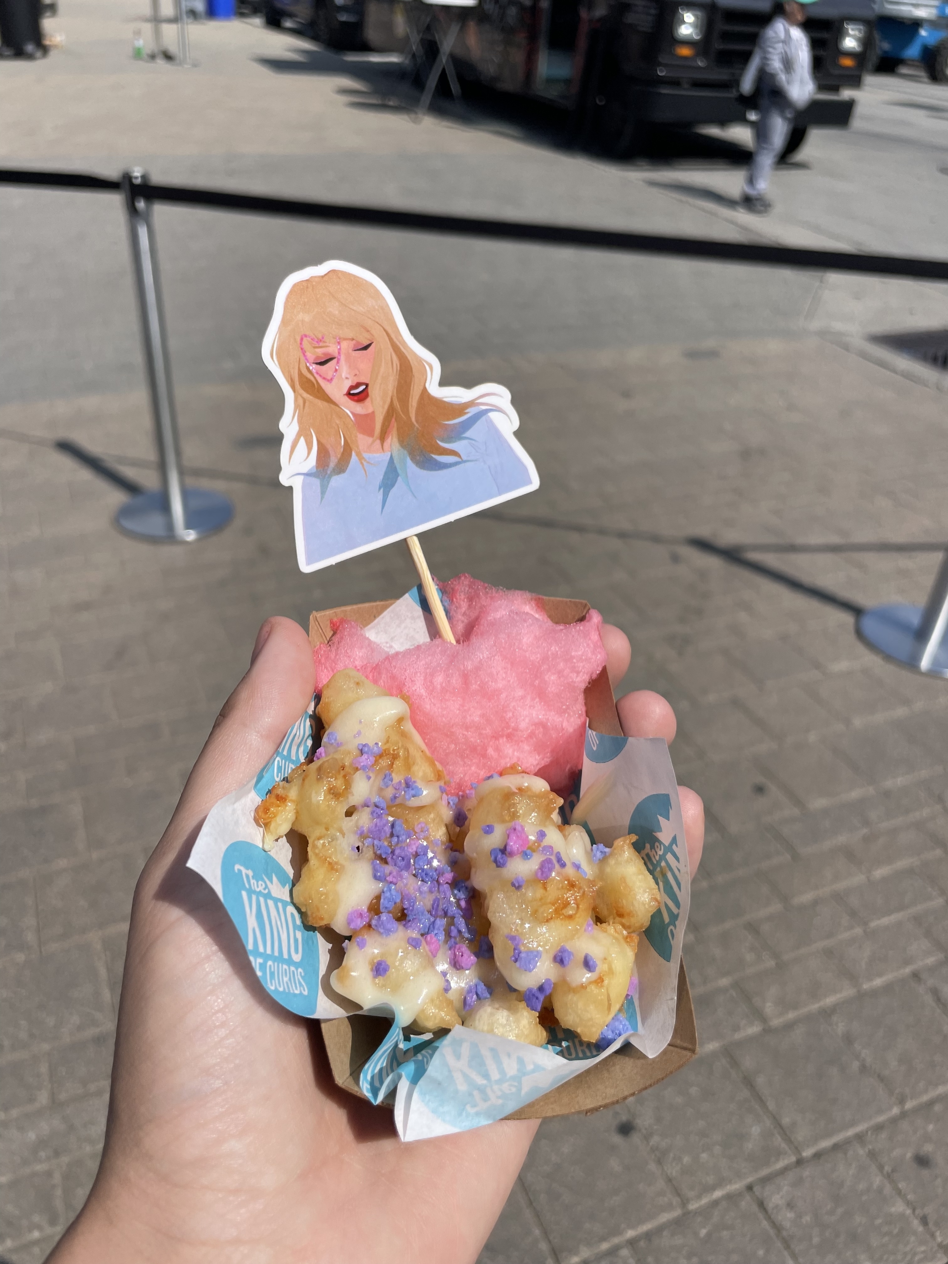 Hand holding a dish with tater tots topped with cheese and purple sprinkles, pink cotton candy, and a stick with a cartoon woman on it. Background shows a food truck