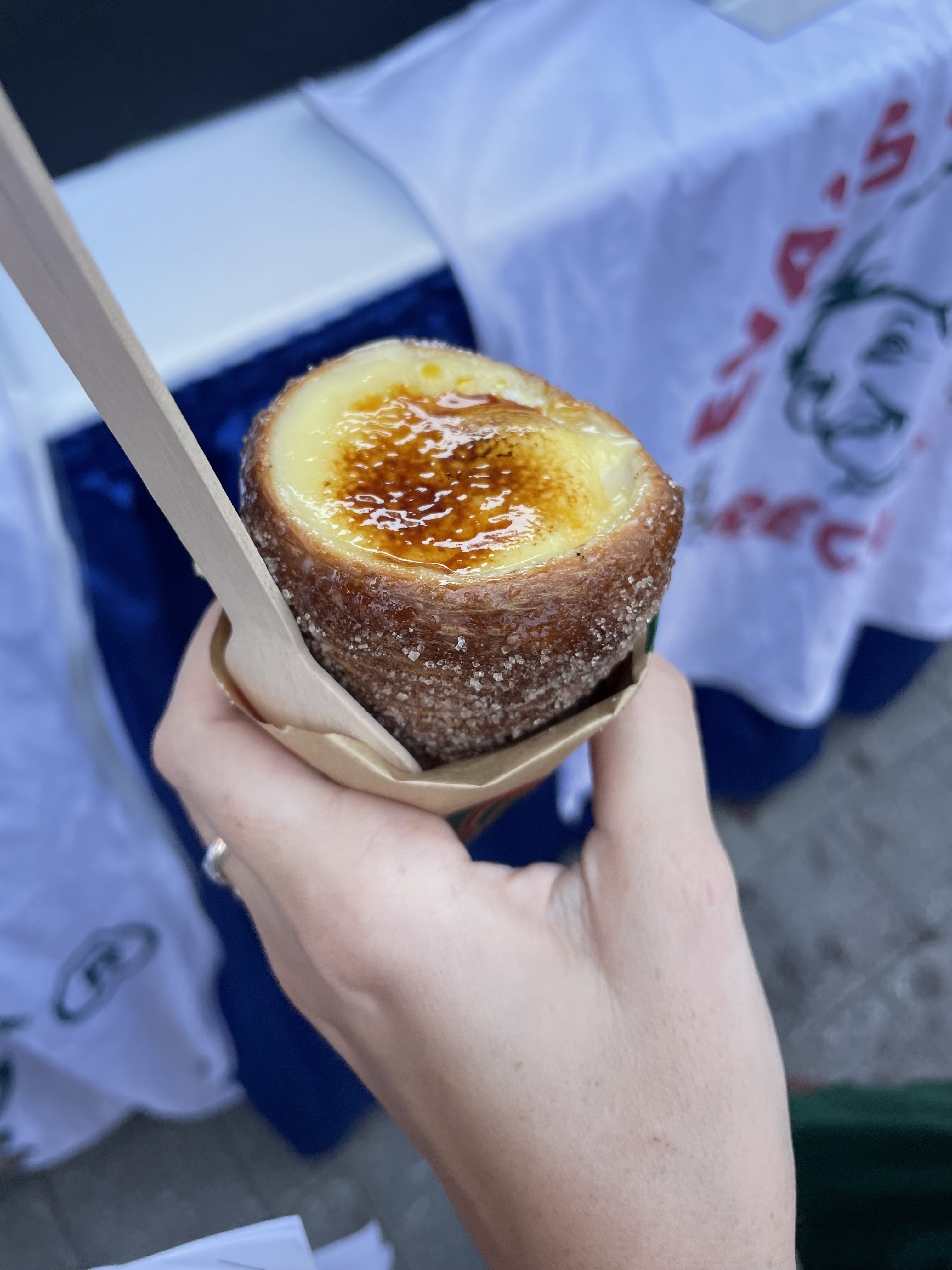 Hand holding a cronut filled with custard, topped with caramelized sugar, and a wooden spoon inserted in it, at a food event or market