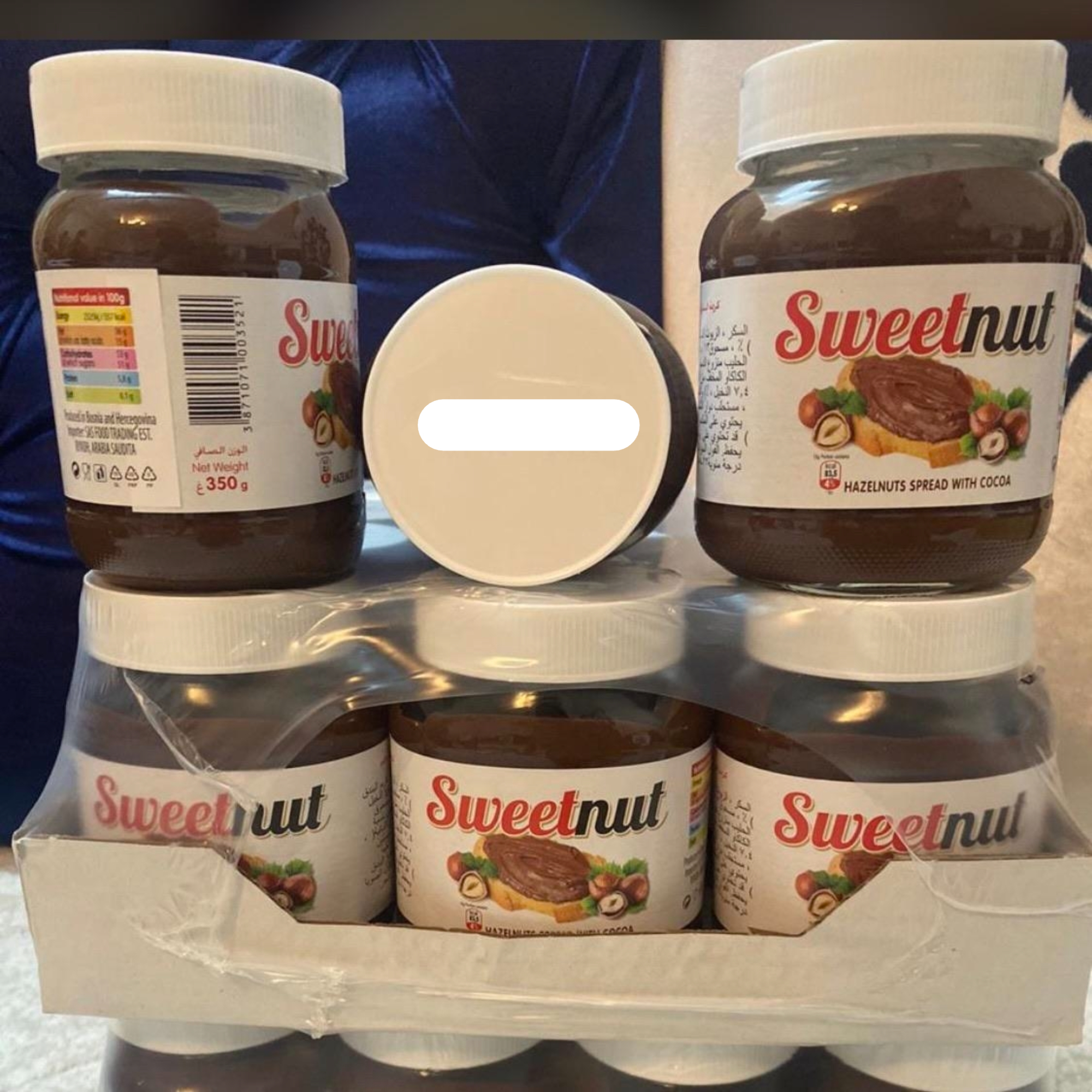 Six jars of Sweetnut hazelnut spread with cocoa stacked, with the expiration date "10/02/2021" visible on the top. The labels show nutritional information and ingredients