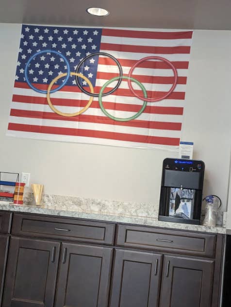An Olympic flag with rings on a U.S. flag background hangs above a countertop with a water dispenser and paper cups