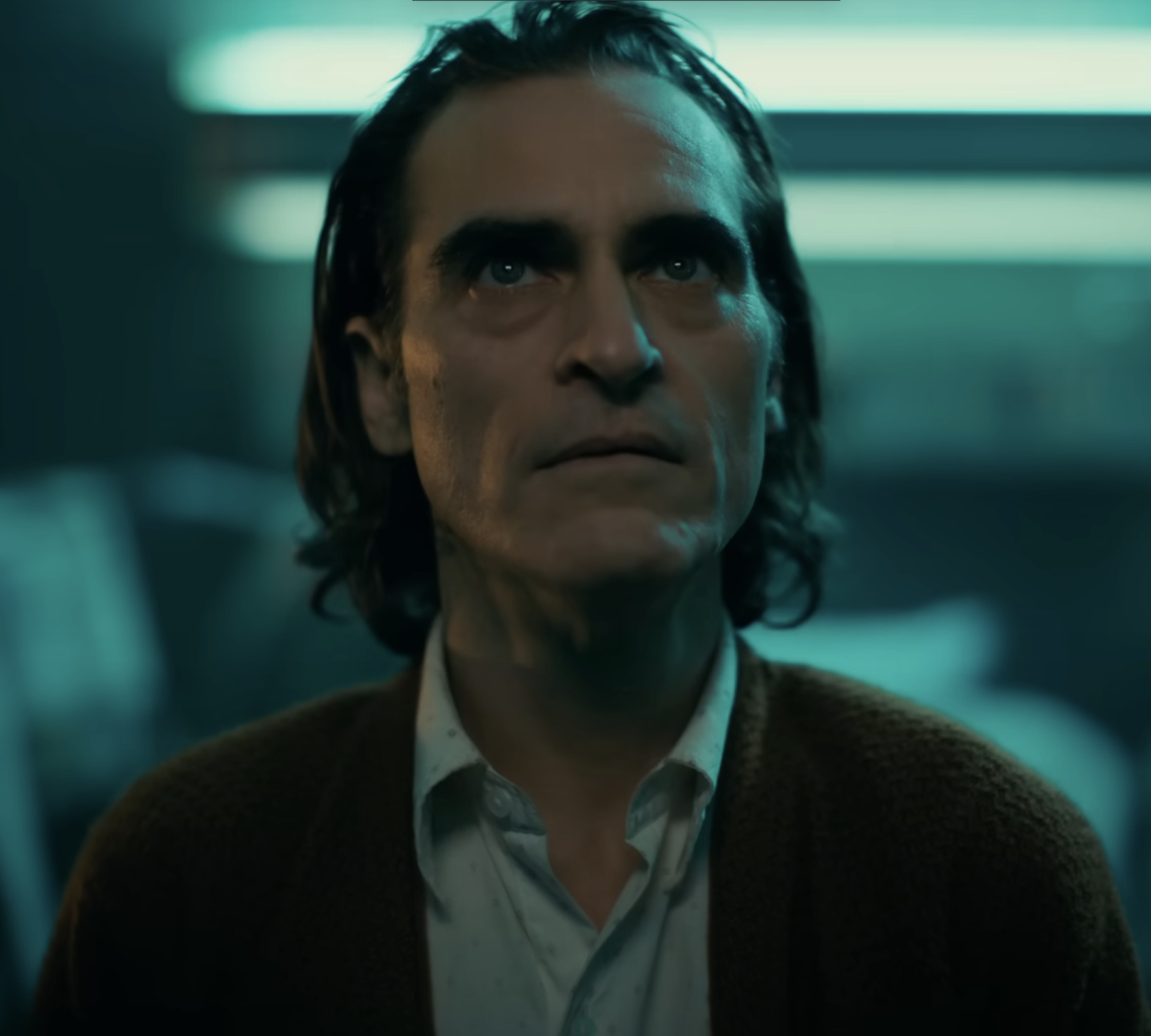 Joaquin Phoenix, in a scene from the movie "Joker," looking upward with a serious expression, wearing a button-up shirt and a cardigan sweater