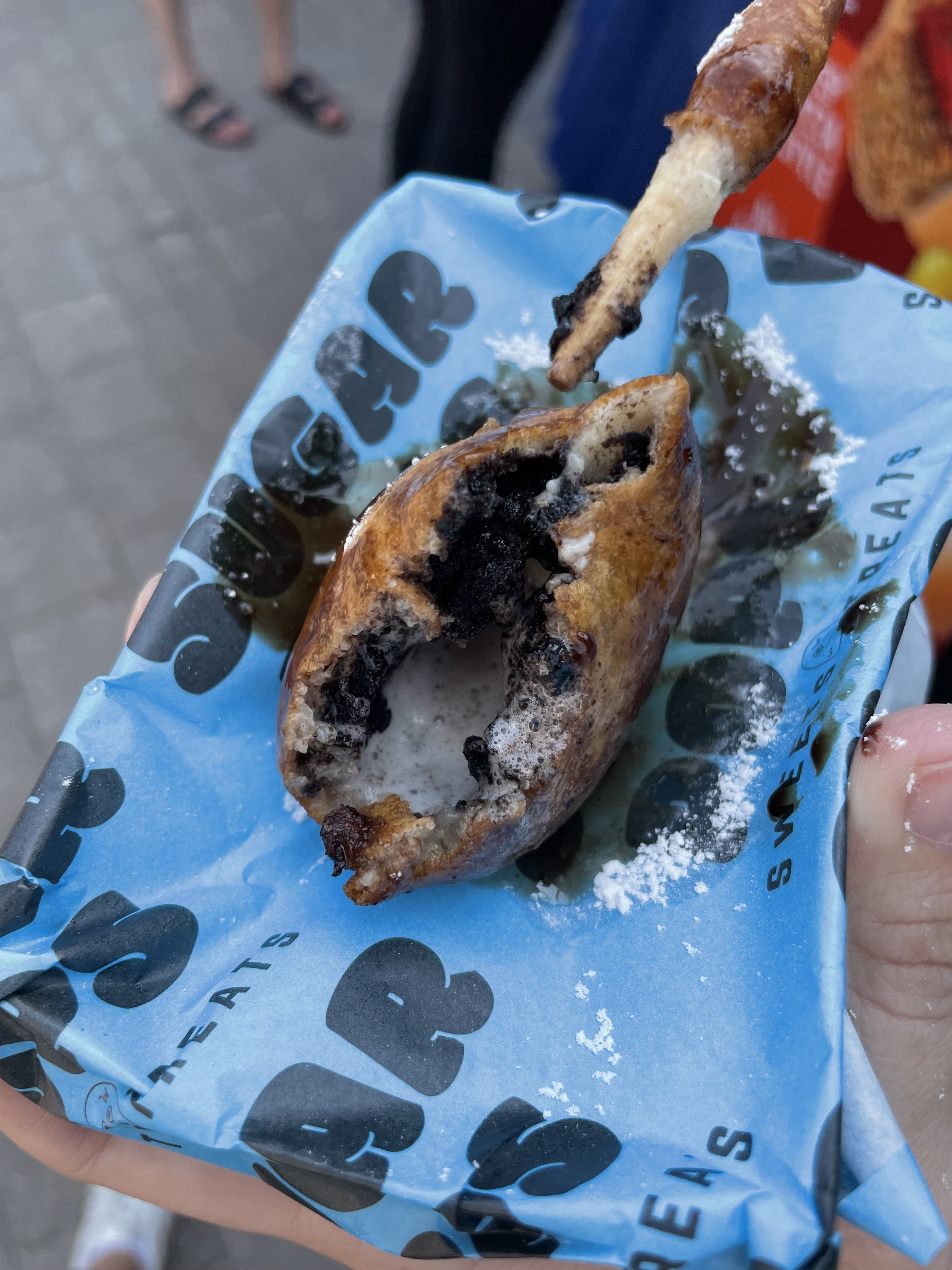 Close-up of a deep-fried Oreo served on a napkin, with someone holding a fried stick in the background