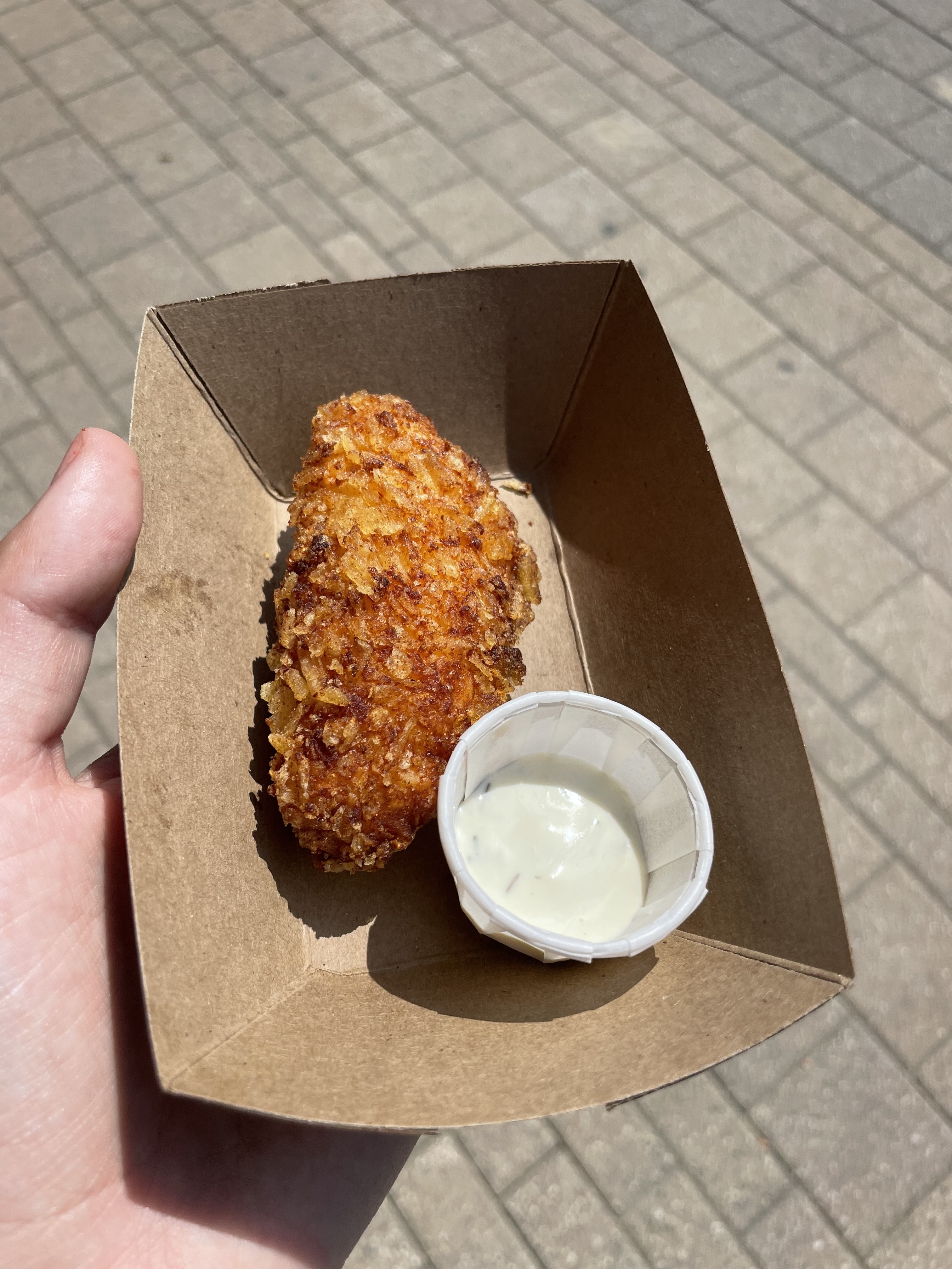A hand holding a cardboard tray with a single breaded and fried food item next to a small cup of white dipping sauce