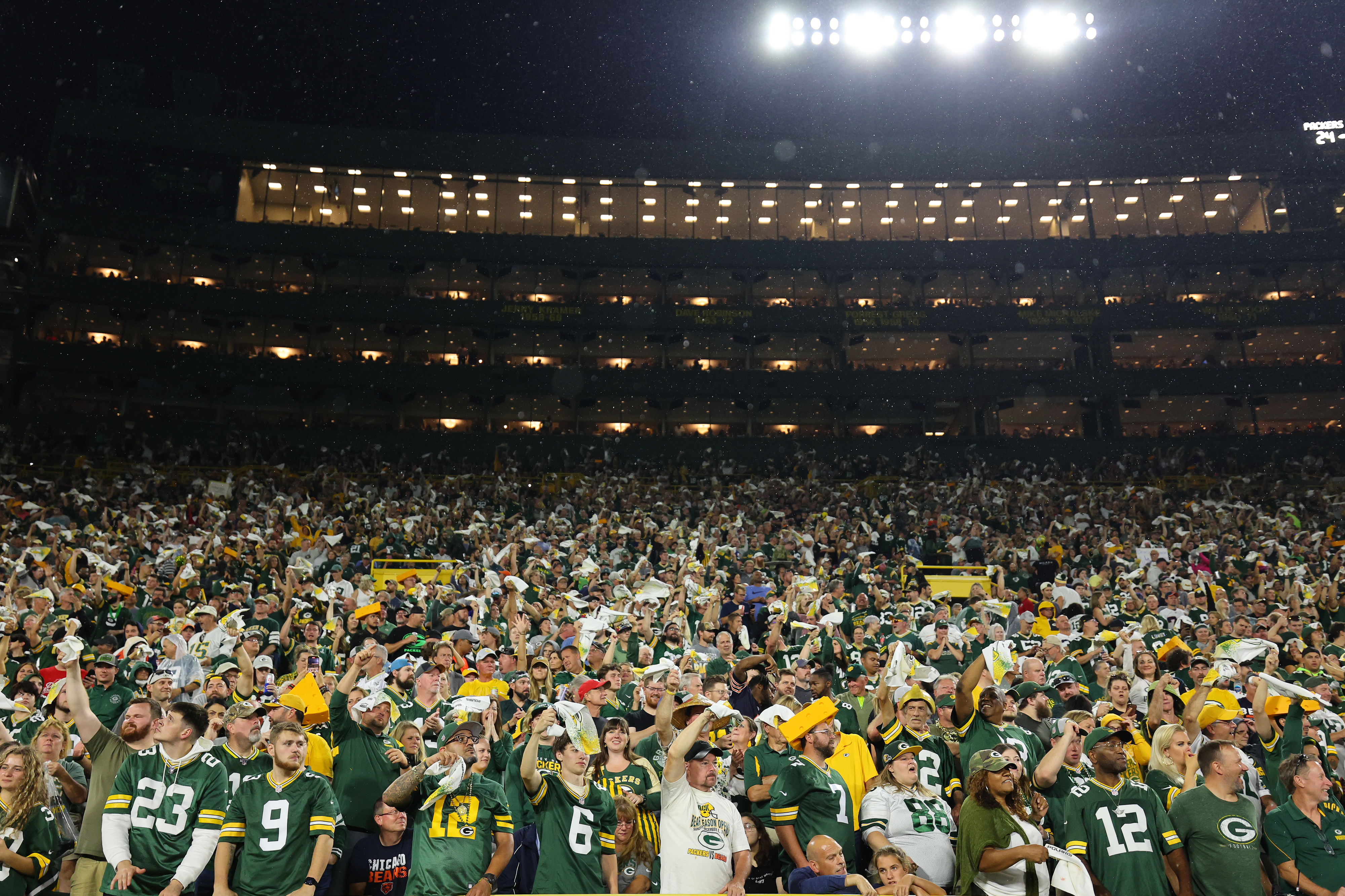 A large crowd of Green Bay Packers fans at a football game, many wearing team jerseys, cheer and wave white towels in a brightly lit stadium