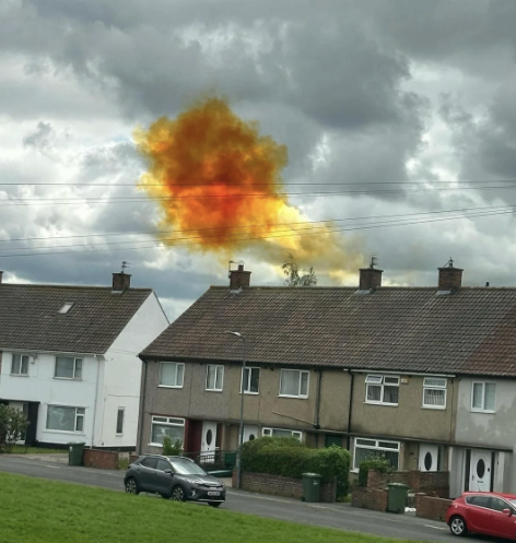 A fiery orange explosion appears in the sky above a row of houses in a suburban neighborhood with a cloudy backdrop