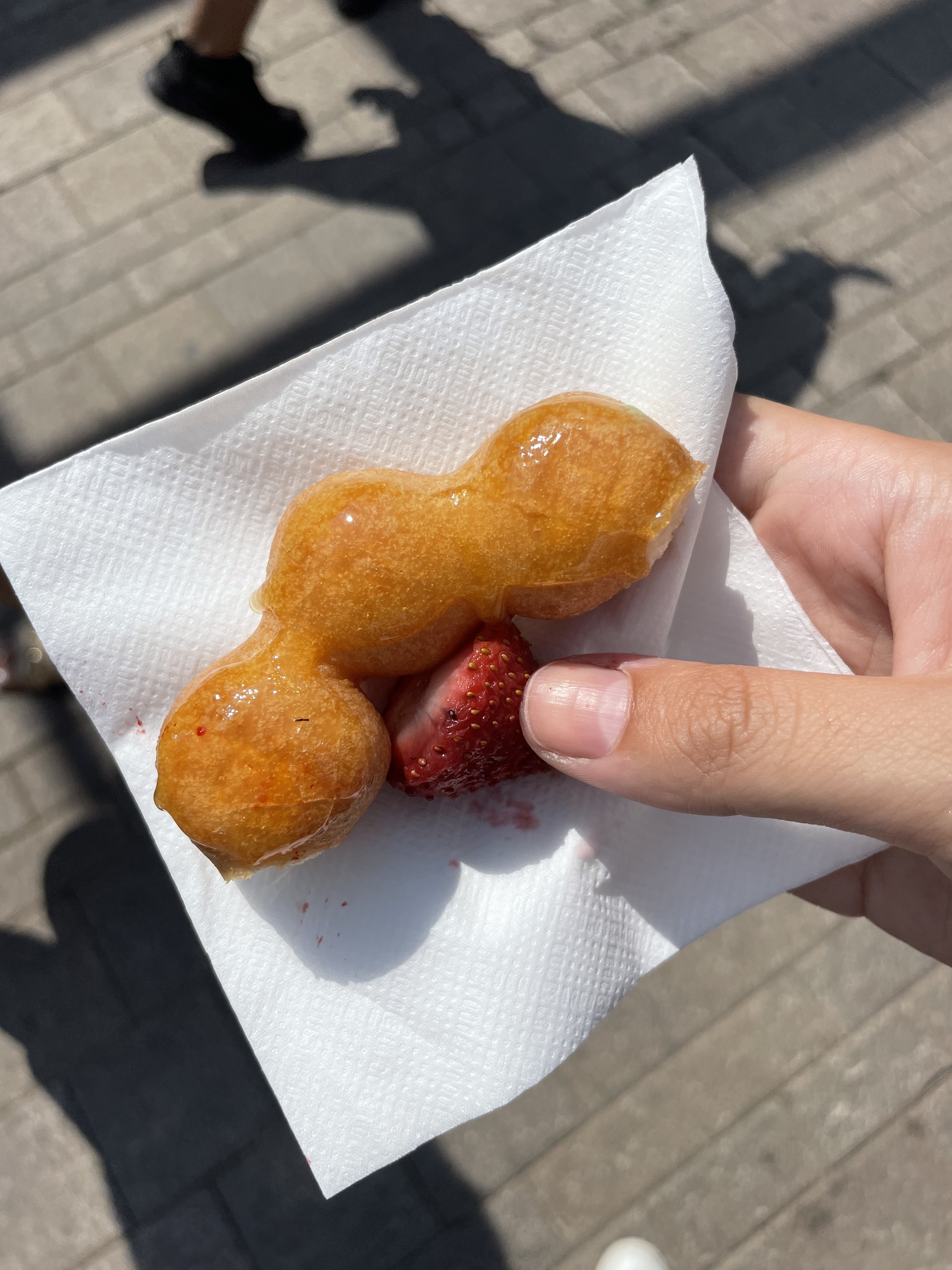 Hand holding a napkin with a small, uniquely shaped corn dog and a half-eaten strawberry, outdoors with people walking in the background