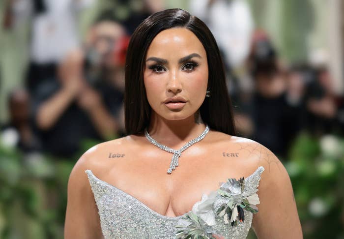 Demi Lovato in an elegant, off-shoulder gown adorned with floral details at a public event, wearing a diamond necklace