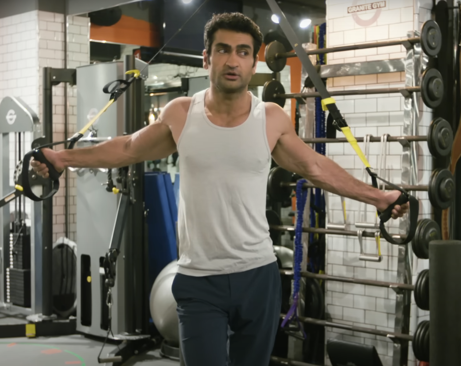 Kumail Nanjiani works out in a gym, using resistance bands for arm exercises. He wears a sleeveless top and athletic pants