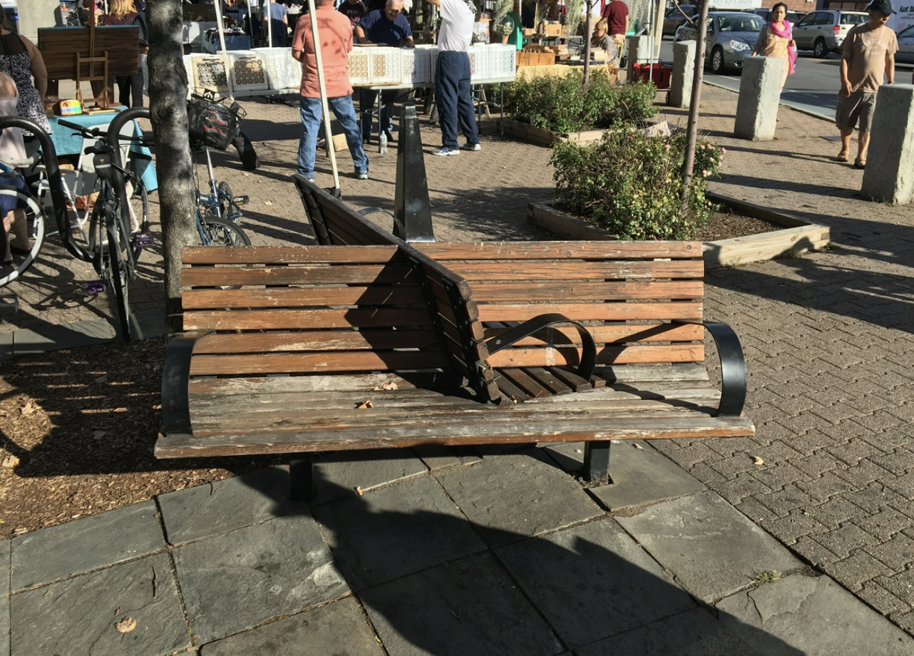 Park bench with a raised metal bar preventing sitting, located in a busy outdoor area with people, tables, and market stalls in the background