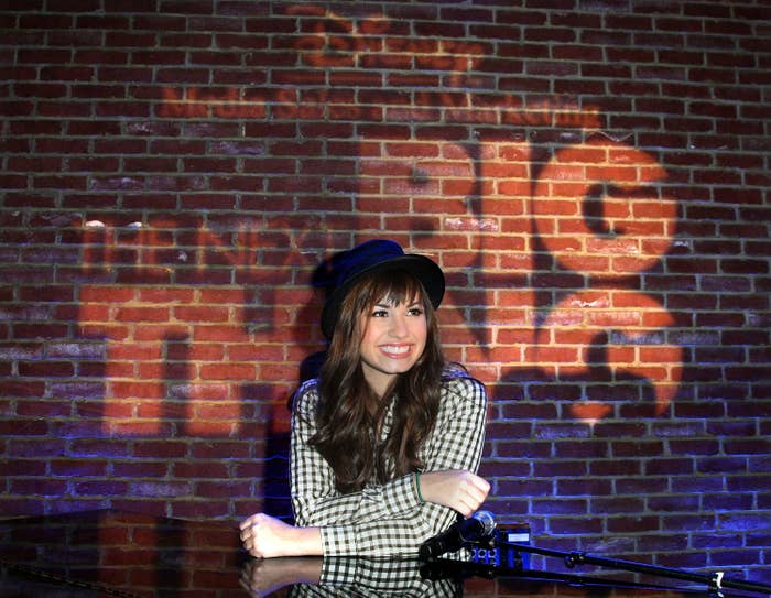 Demi Lovato sits behind a piano, smiling, wearing a hat and checkered shirt, with a brick wall as the backdrop