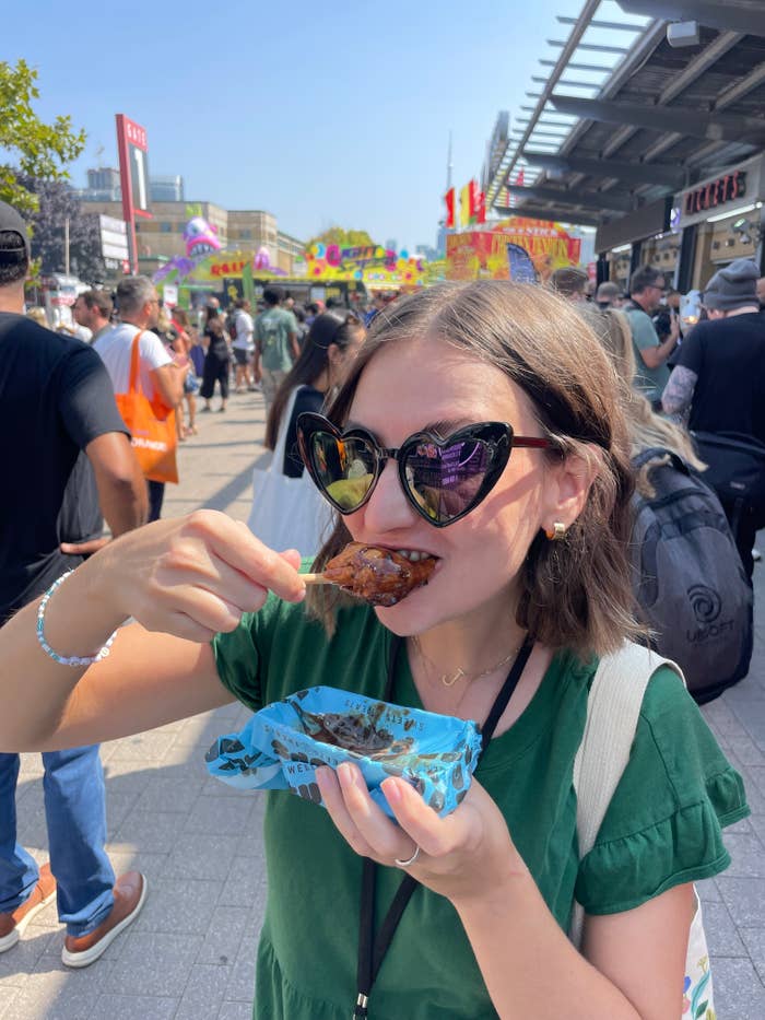 Woman enjoys a chicken wing at a busy outdoor food festival, holding a blue paper tray. She wears large black cat-eye sunglasses and a green short-sleeve top