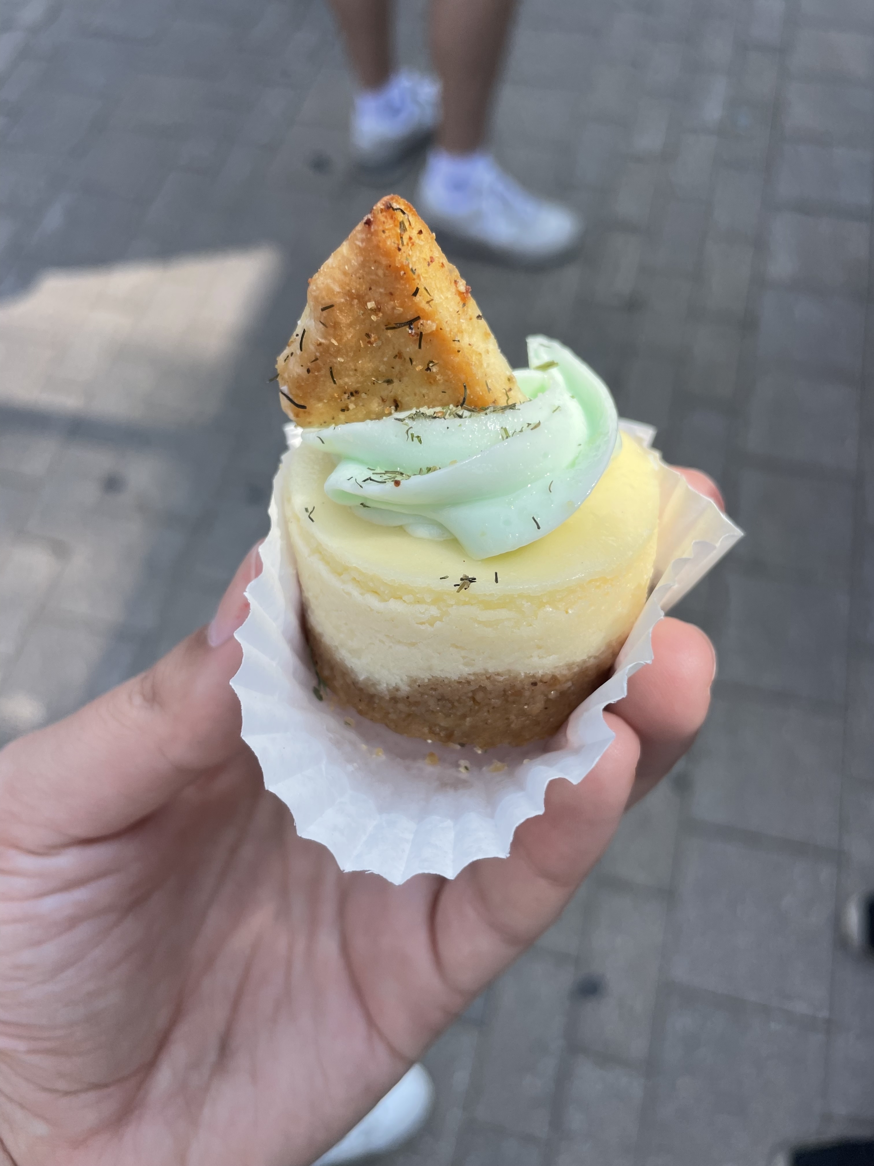 Close-up of a hand holding a mini cheesecake topped with green frosting and a small pastry triangle garnish. People’s legs are visible in the background