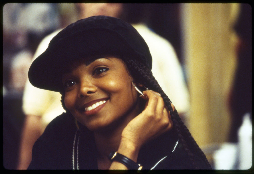 Janet Jackson smiling, wearing a black hat and hoop earrings, resting her chin on her hand