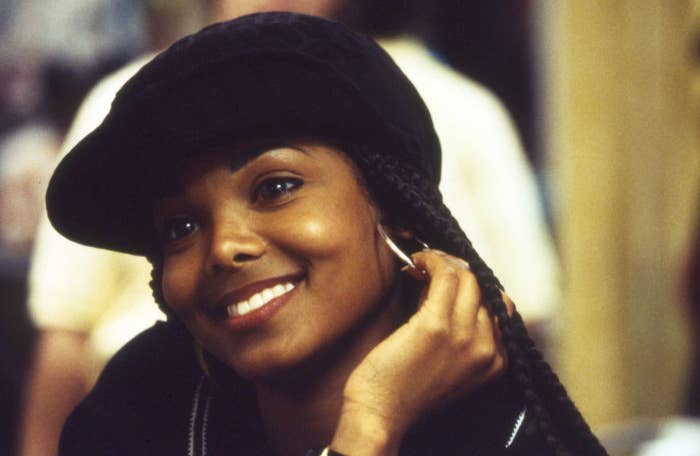 Janet Jackson smiling, wearing a black hat and hoop earrings, resting her chin on her hand