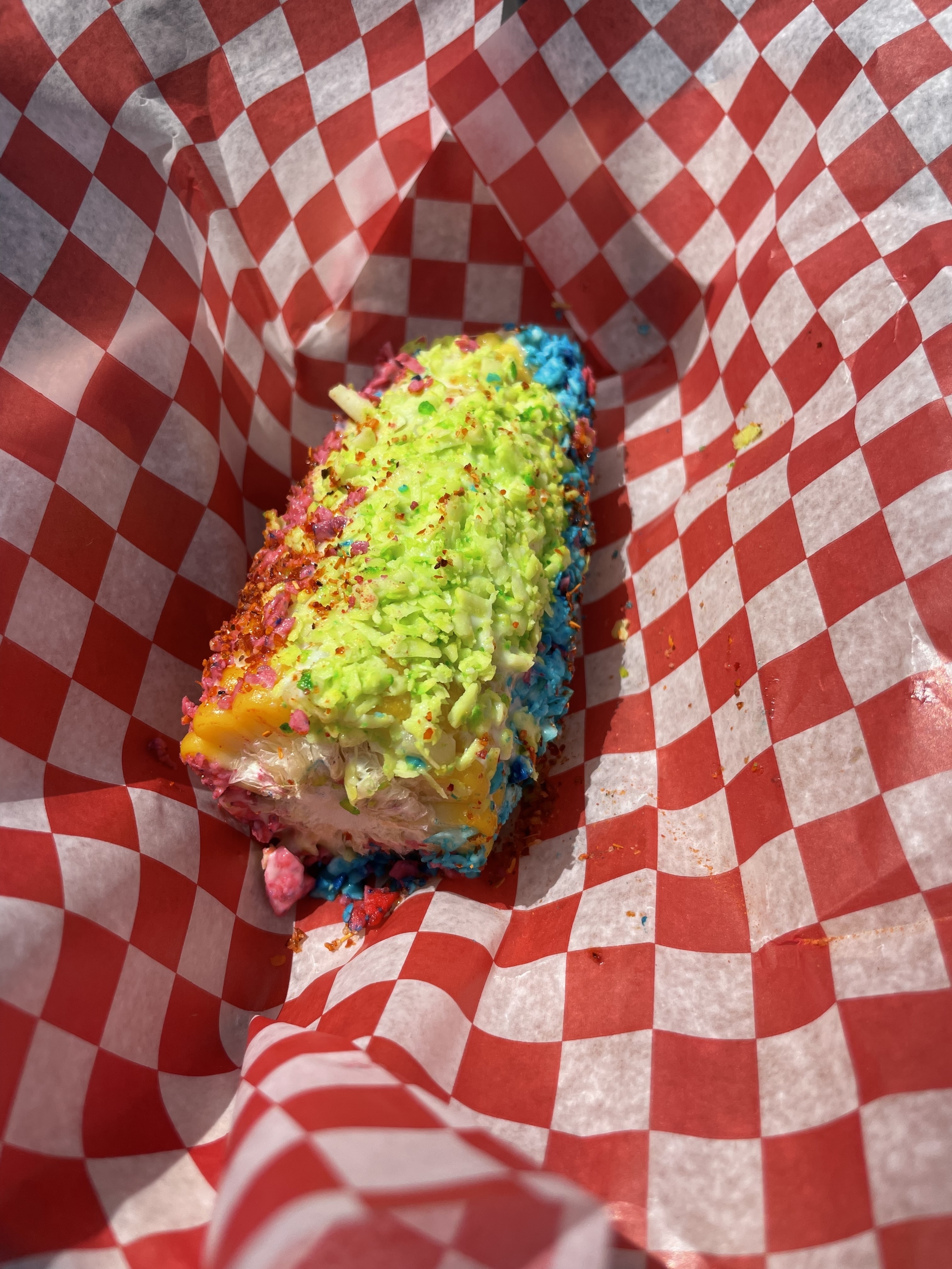 A brightly colored, coated corn on the cob is placed on a red and white checkered paper