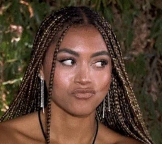 A woman with long braids and silver earrings has a playful smirk and looks to the side