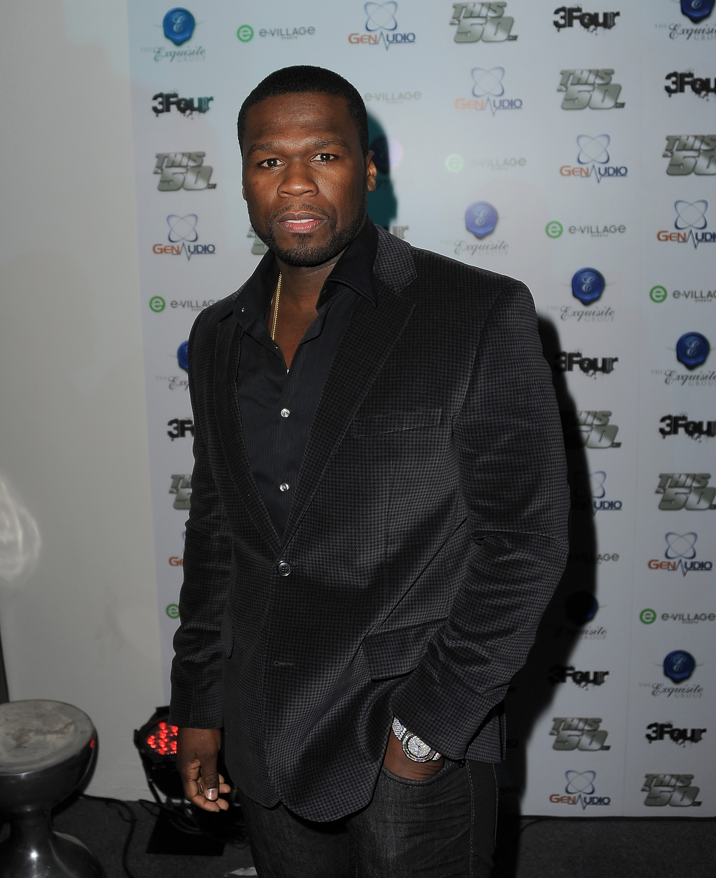 50 Cent in a black suit and shirt poses in front of a branded backdrop at an event