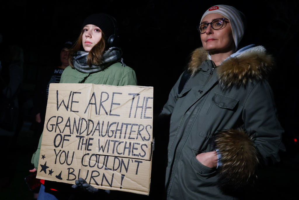 Two people in winter coats hold a sign reading "We are the granddaughters of the witches you couldn't burn!"
