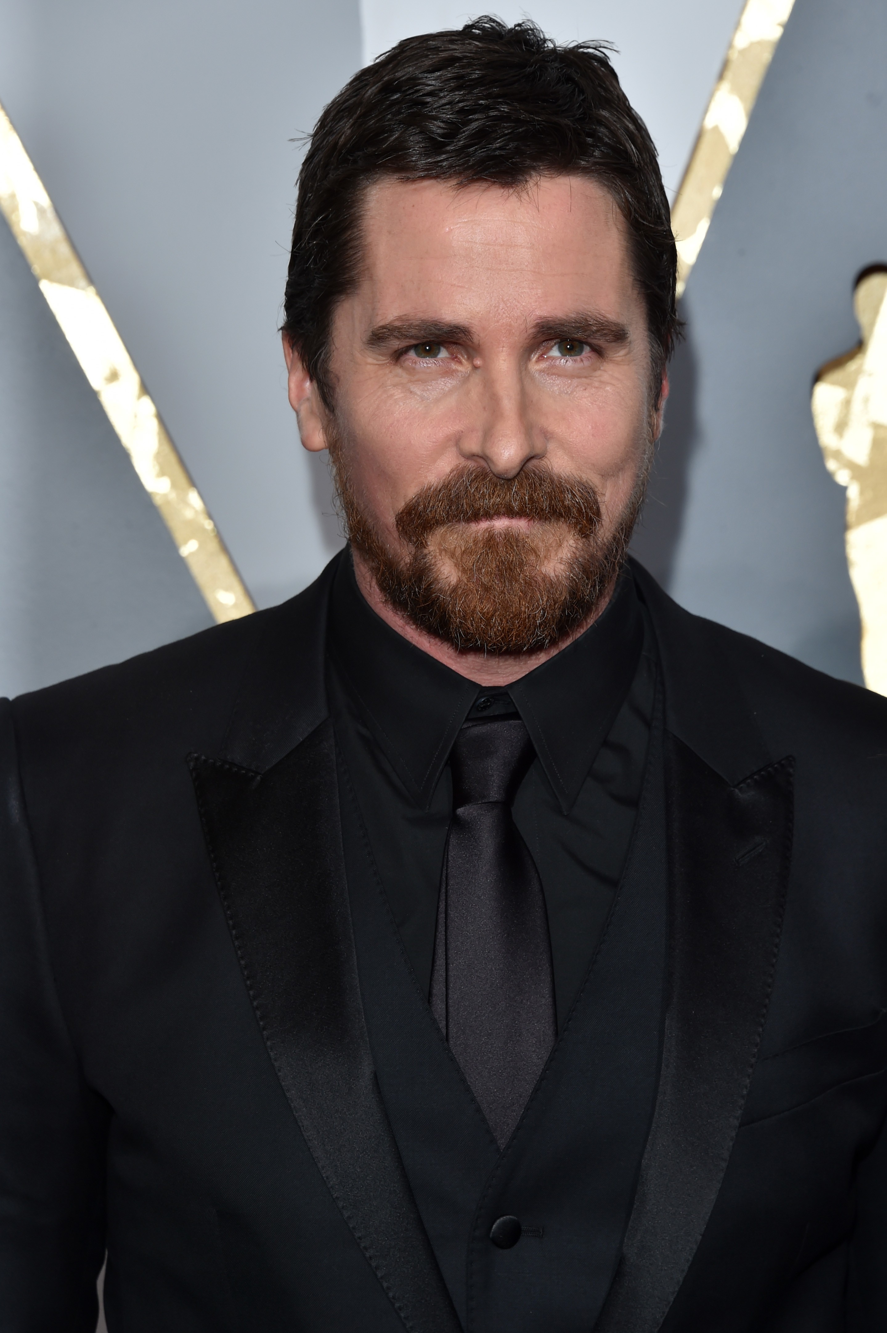 Christian Bale attending an event in a black suit and tie, with a serious expression