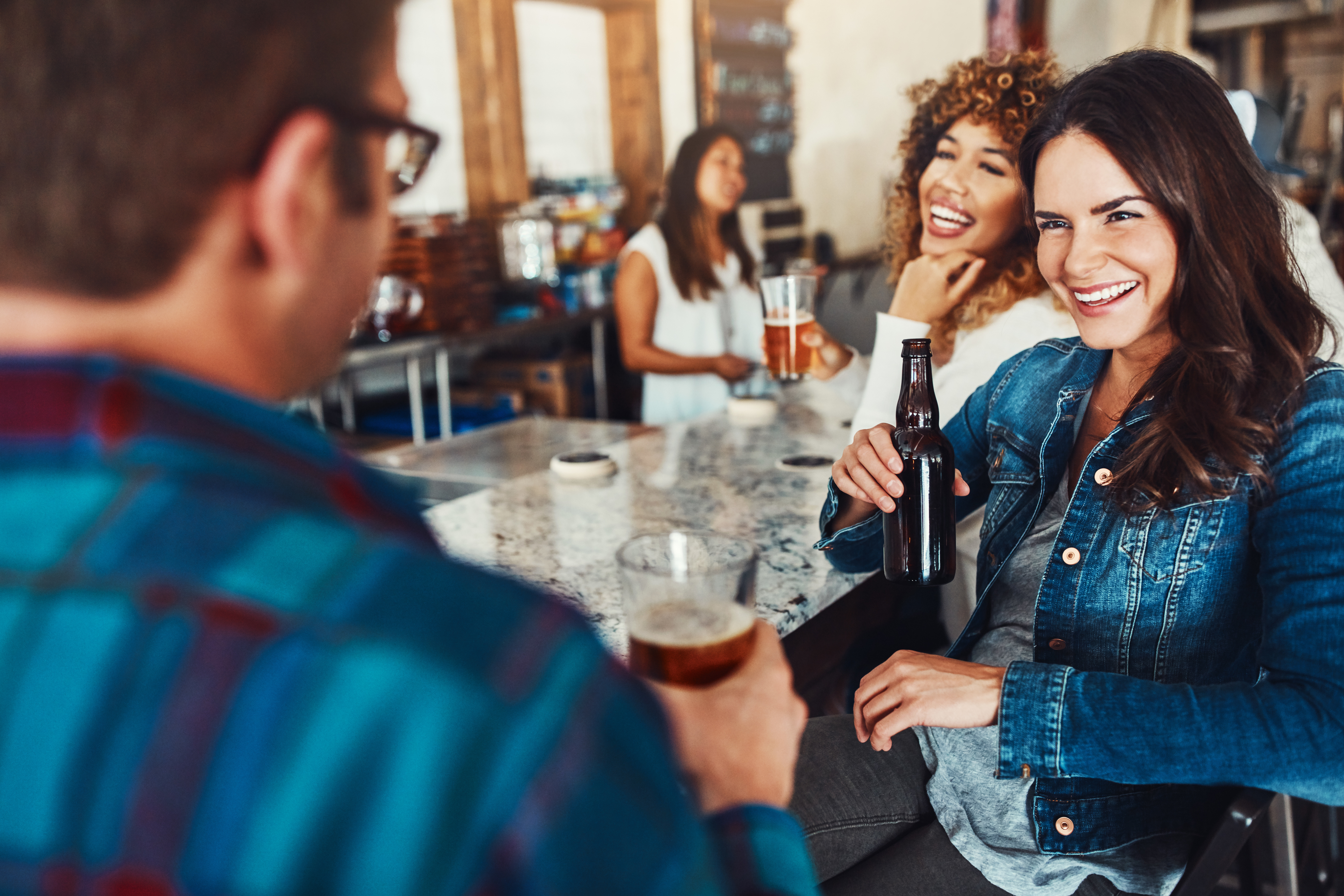 People enjoying drinks at a casual gathering. A woman in a denim jacket smiles while holding a bottle. Other people are chatting in the background