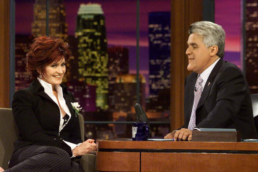 Sharon Osbourne sits and smiles on a talk show set while wearing blazer and blouse. Jay Leno, also in a suit, interviews her