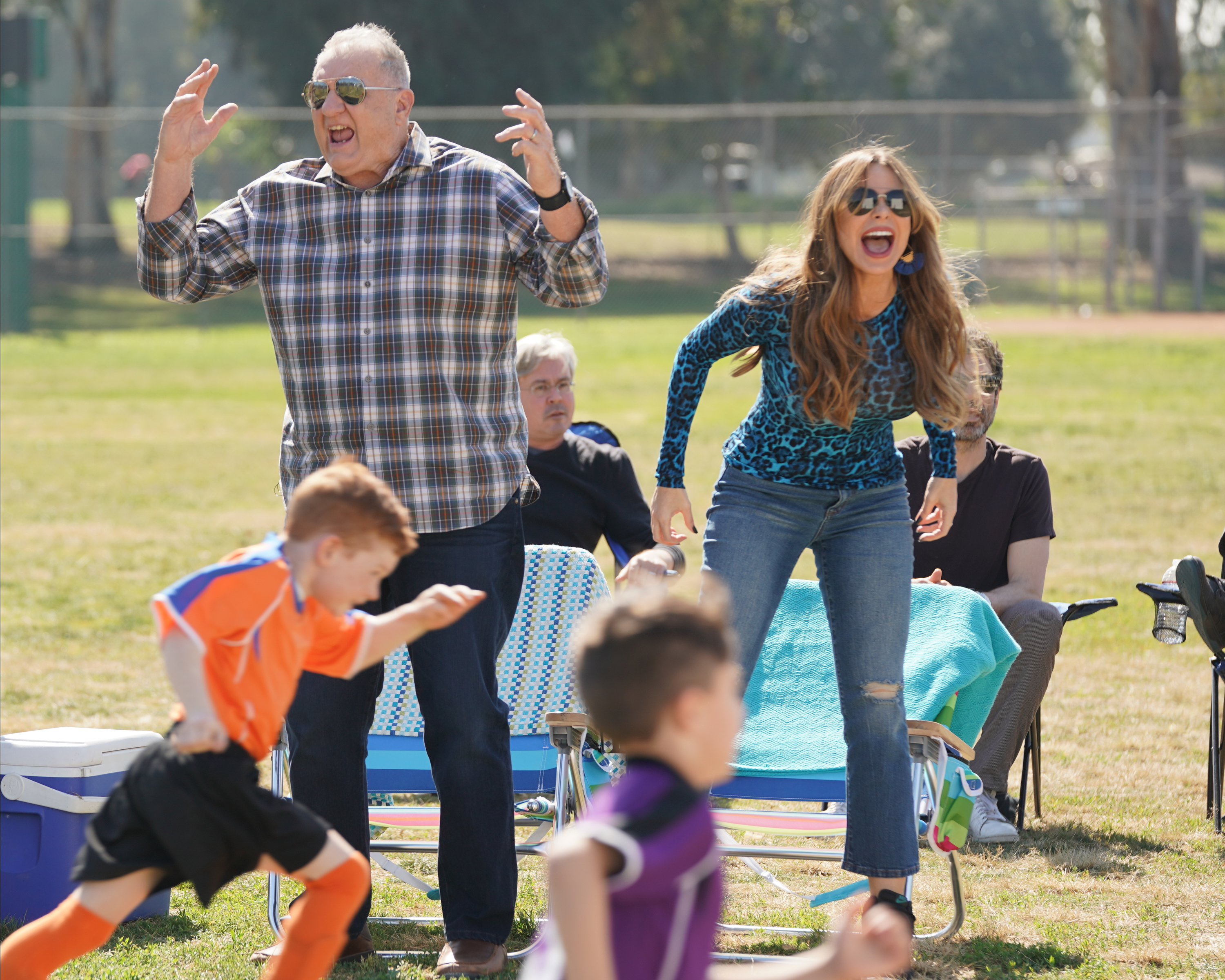 Ed O'Neill and Sofía Vergara enthusiastically cheer from the sidelines of a youth soccer game in the park, in a scene from "Modern Family."