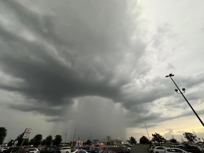 Dark storm clouds loom over a parking lot, with heavy rainfall in the distance. Rows of parked cars and streetlights are visible