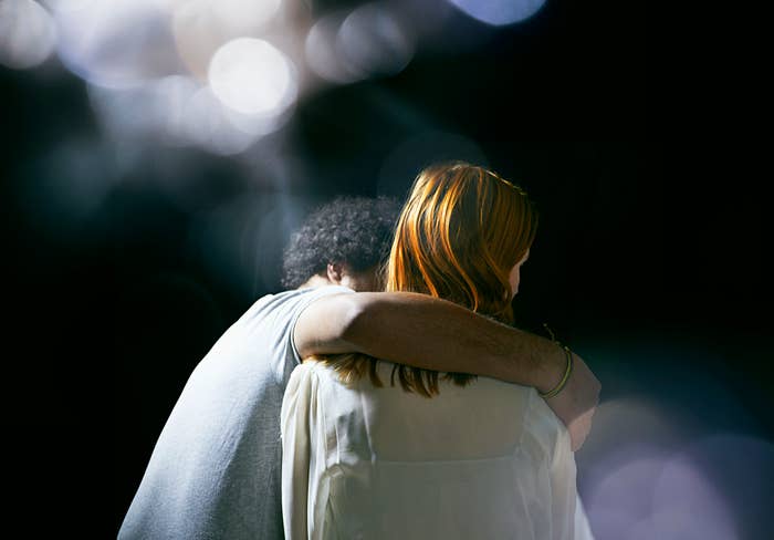 A man with curly hair and a woman with straight hair sit closely with their backs to the camera, the man's arm around the woman's shoulder