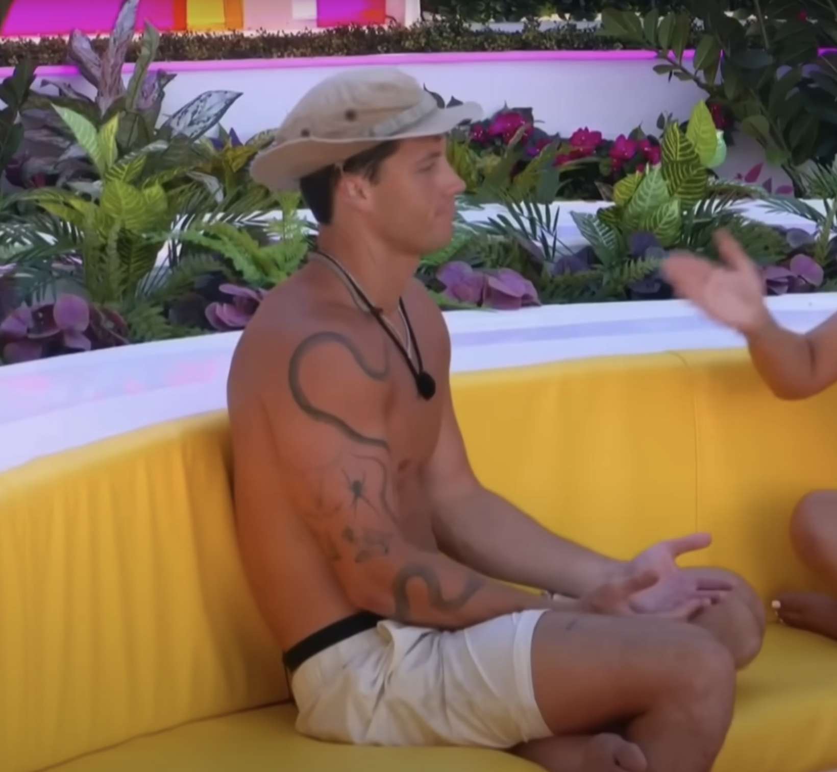 A man in khaki shorts and a safari hat sits shirtless on a yellow bench, gesturing with his hands during a conversation