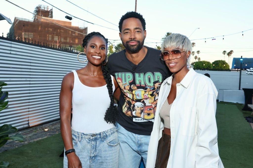 Three individuals, Issa Rae, Jay Ellis, and Yvonne Orji, are smiling while posing together outdoors; Jay Ellis is wearing a graphic T-shirt