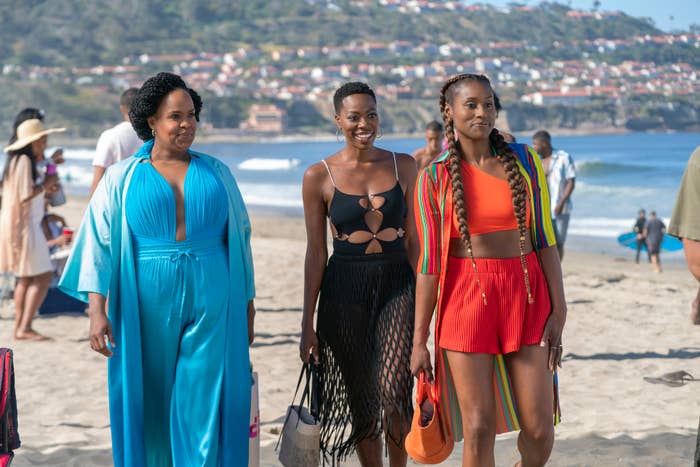 Three women, including Issa Rae (right), walk along a beach smiling. The woman on the left wears a flowing robe, the middle woman a black cut-out dress, and Issa Rae wears a red outfit