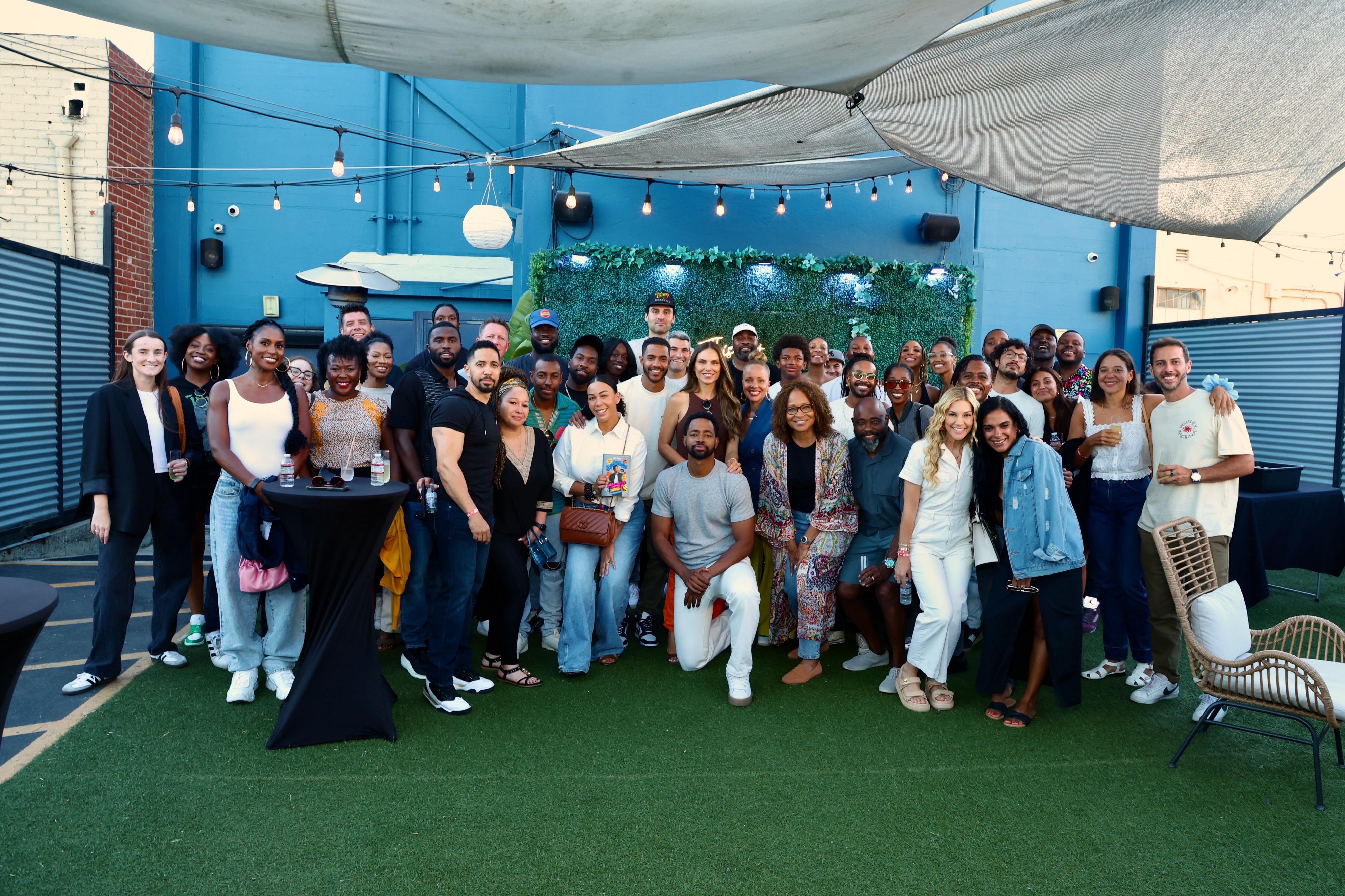 Group of people smiling and posing outdoors, some standing and others crouching, near a decorated wall on a rooftop. Names not provided
