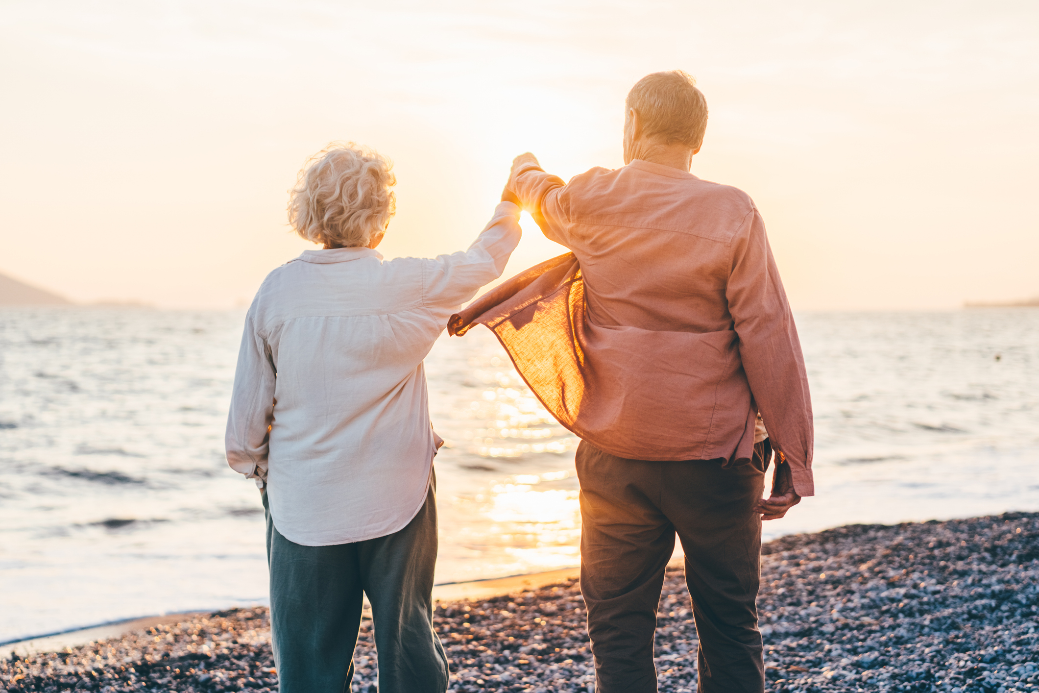 An elderly couple, holding hands on a beach, facing the ocean during sunset, with the man gently pulling the woman's arm up in a playful manner