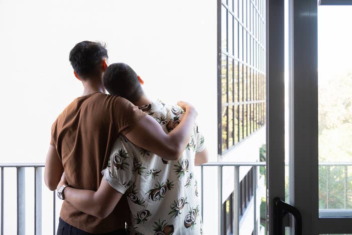 Two people stand on a balcony; one has their arm around the other, with their head resting on their shoulder, showing a moment of comfort and closeness