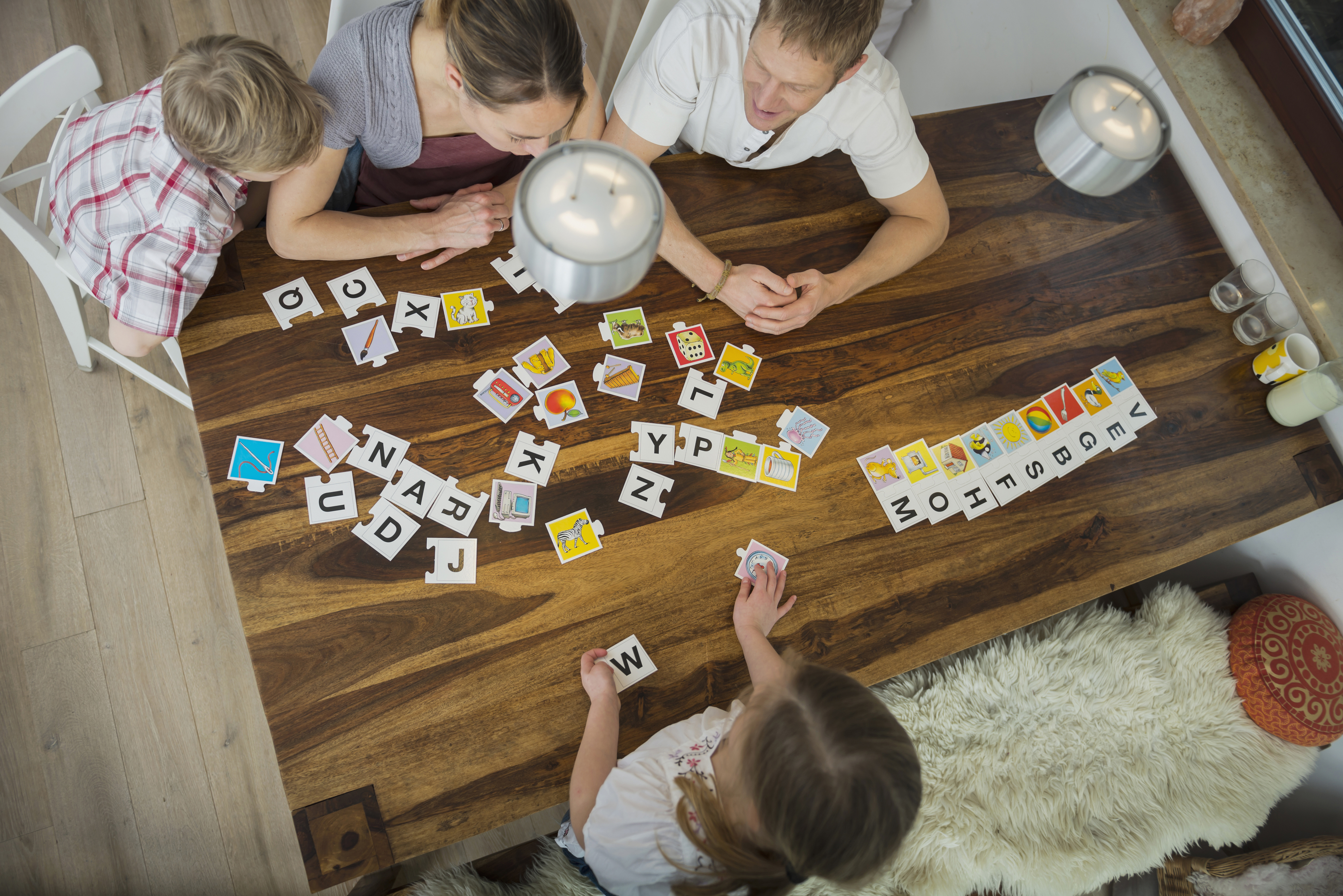 A family of four, two parents and two children, sit around a wooden table playing an educational letter and picture card game