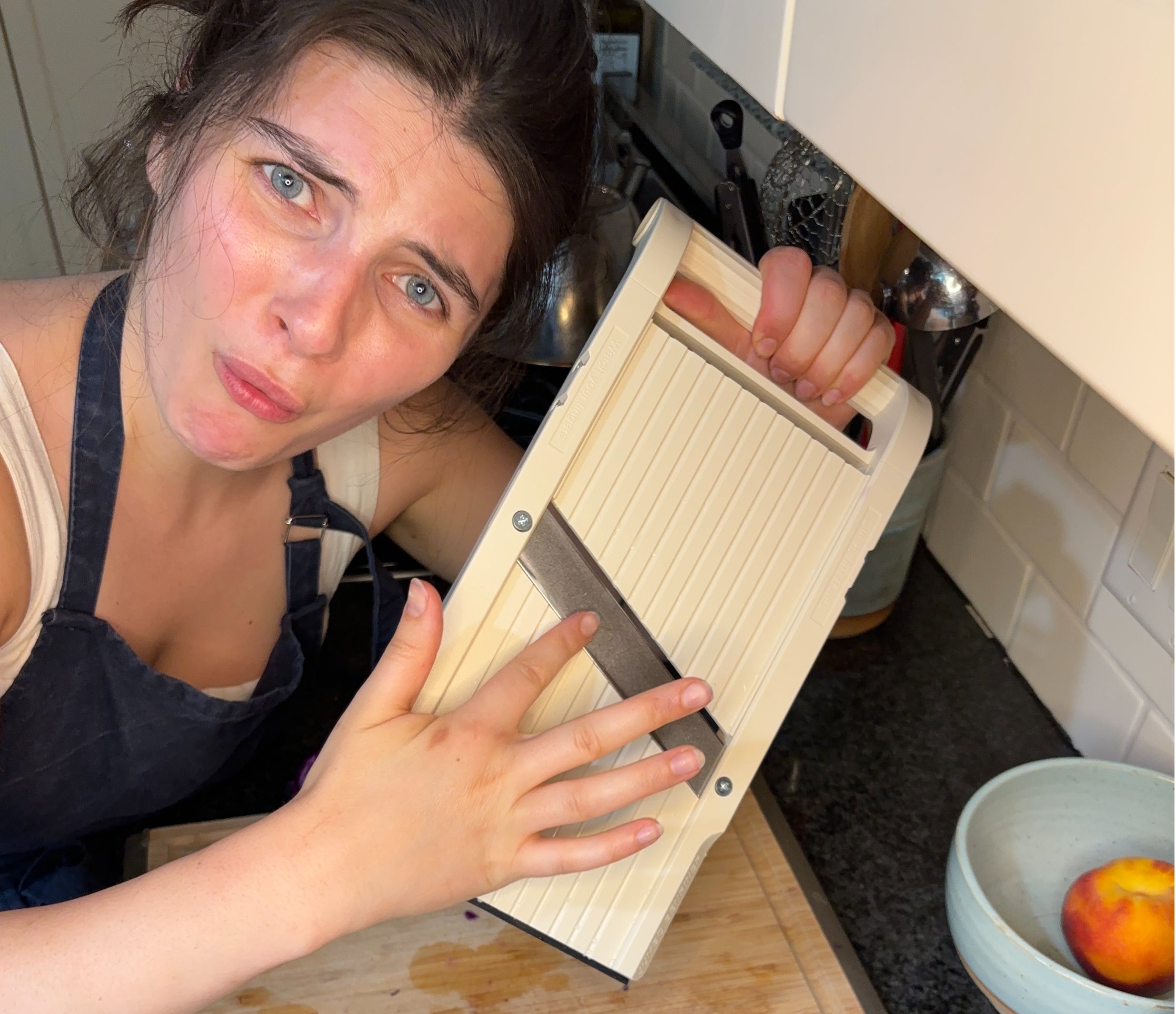 A person in a kitchen is looking confused while holding a mandoline slicer over a cutting board with a piece of cabbage nearby