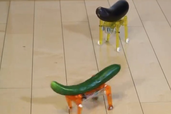 部屋の床に、小さなロボット脚が付いたキュウリとナスの模型が置かれている。