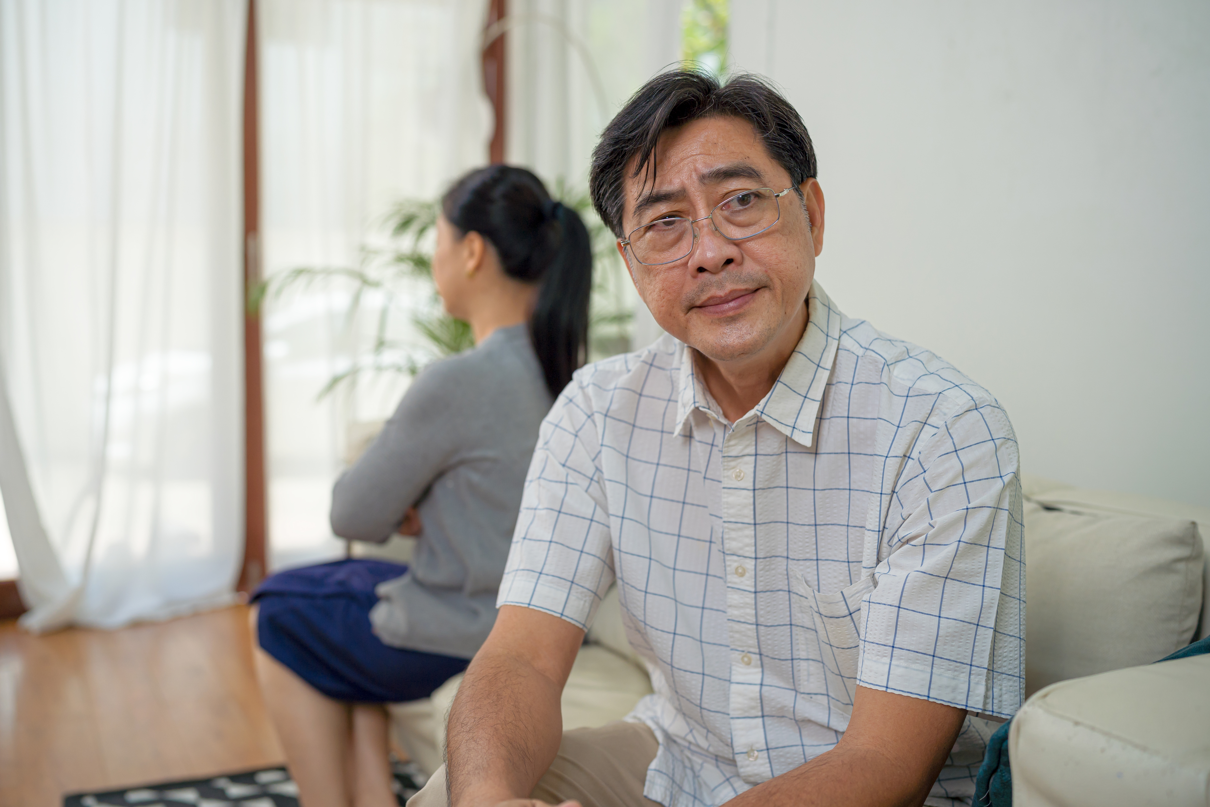 An elderly man with glasses and a checkered shirt sits on a couch, looking at the camera. A woman with her back turned sits beside him