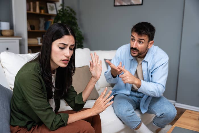 A woman looks upset and raises her hand while a man sitting next to her gestures emphatically during a conversation in a living room setting