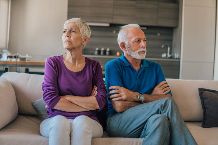 An older woman and man sitting on a couch with arms crossed, both looking unhappy and not facing each other