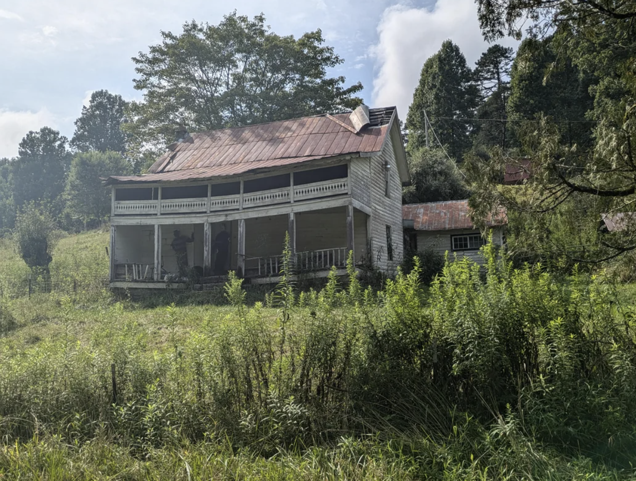 An old, weathered house stands in a lush, overgrown field with tall grass and surrounding trees in the background. The image evokes a sense of abandonment