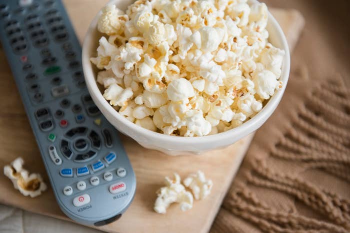 A bowl of popcorn sits next to a remote control on a wooden surface with a knitted fabric in the background