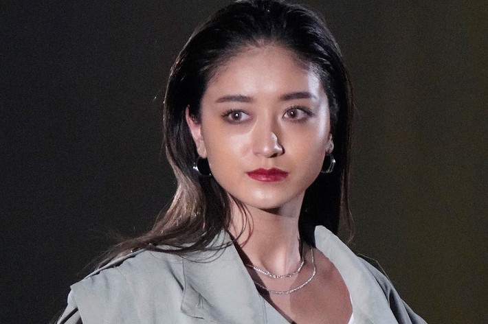 女性モデルが、白いインナーとオーバーサイズのグレーコートを着て、カメラに向かってポーズをとっている。