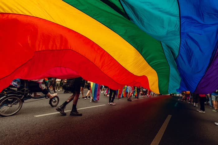 People participating in a parade carrying a large rainbow flag above their heads. Some participants are walking, while one is seen in a wheelchair