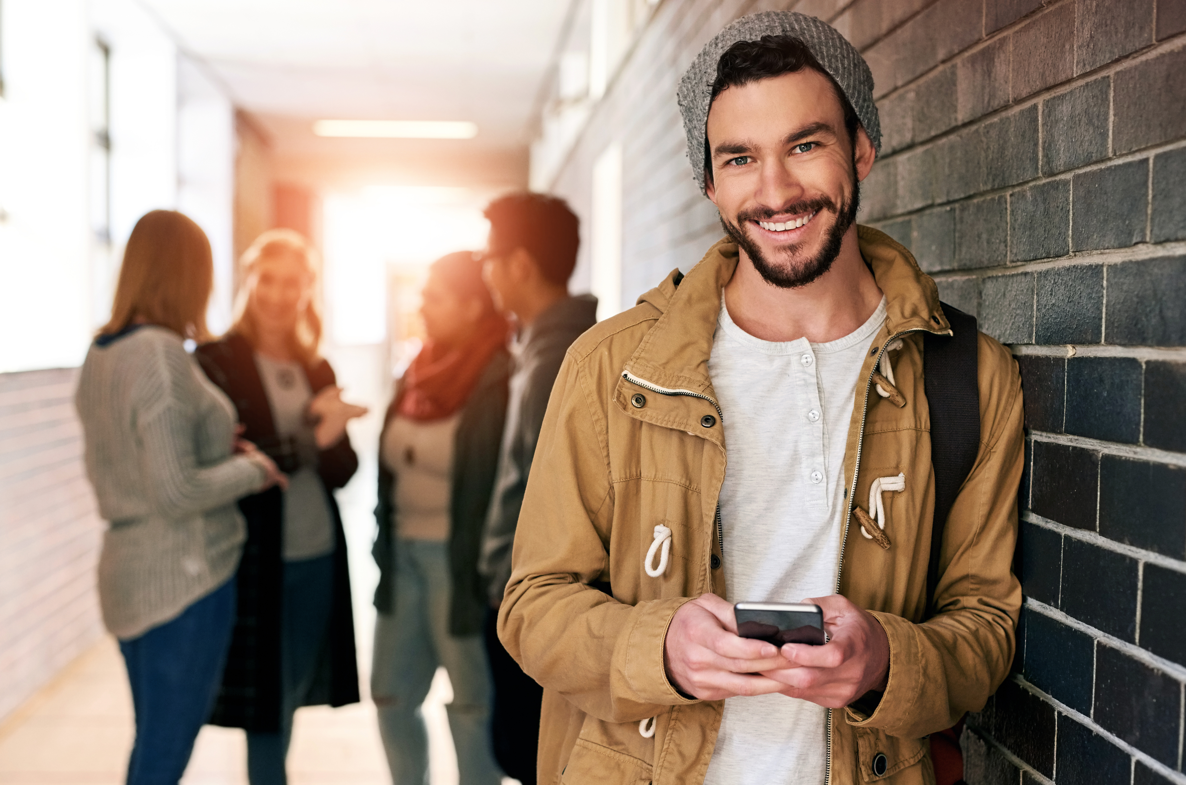 A smiling man in casual clothing with a beanie and jacket leans against a brick wall, holding a smartphone. Four people are conversing in the background. Names unknown