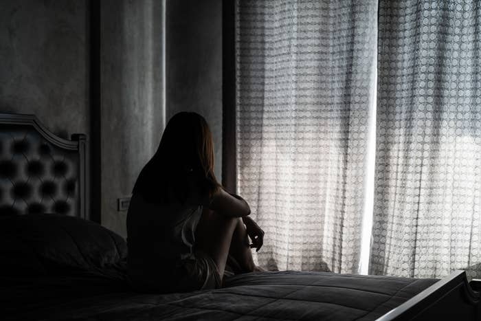 Woman sits in a dark room on a bed, looking out through partially closed curtains