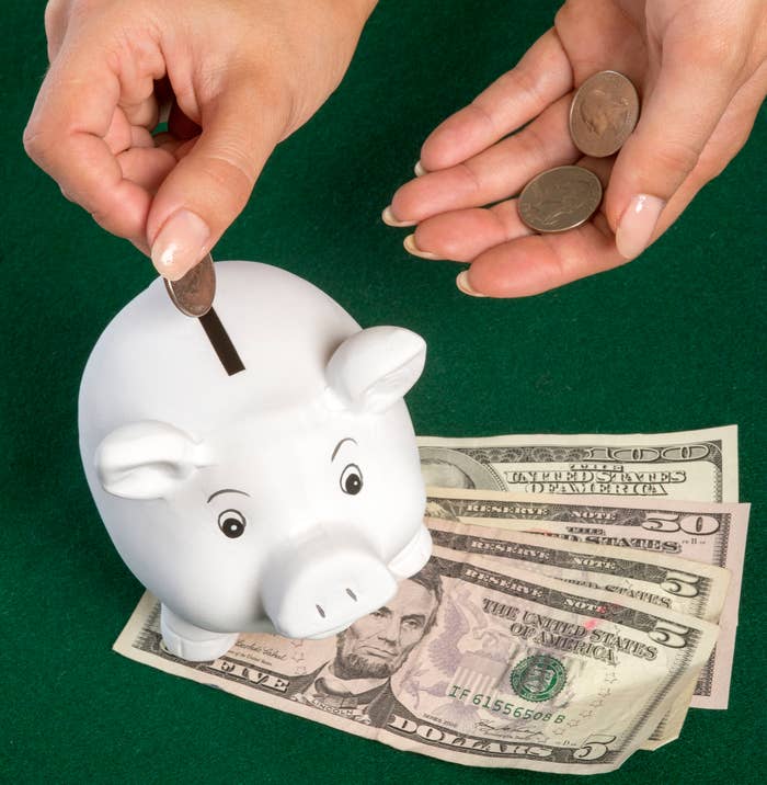 Hands placing coins into a white piggy bank with USD bills on a green surface