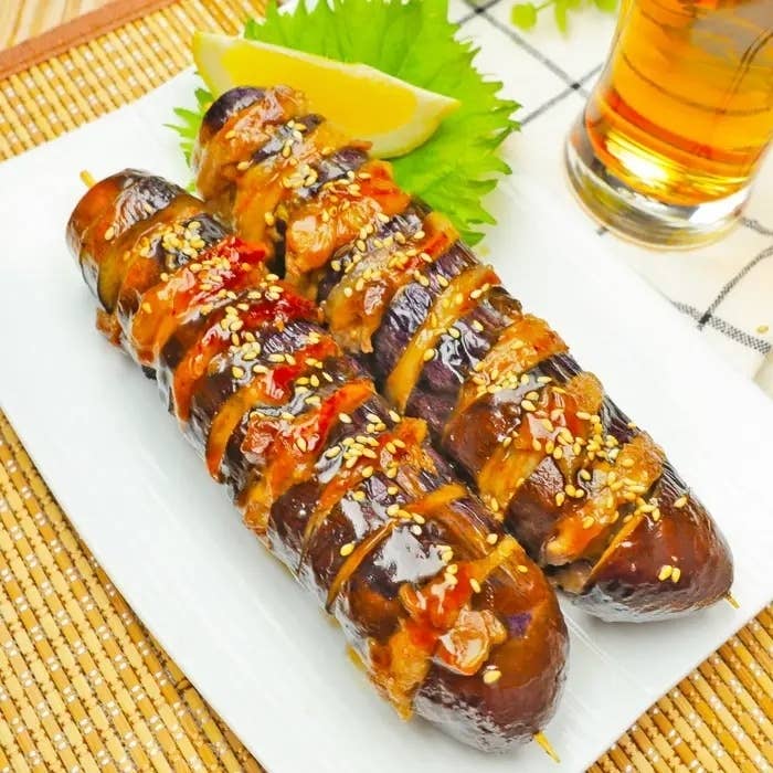 美味しそうな焼き茄子がタレとゴマで飾られて皿に載せられています。すぐ横にグラス入りの飲み物があります。