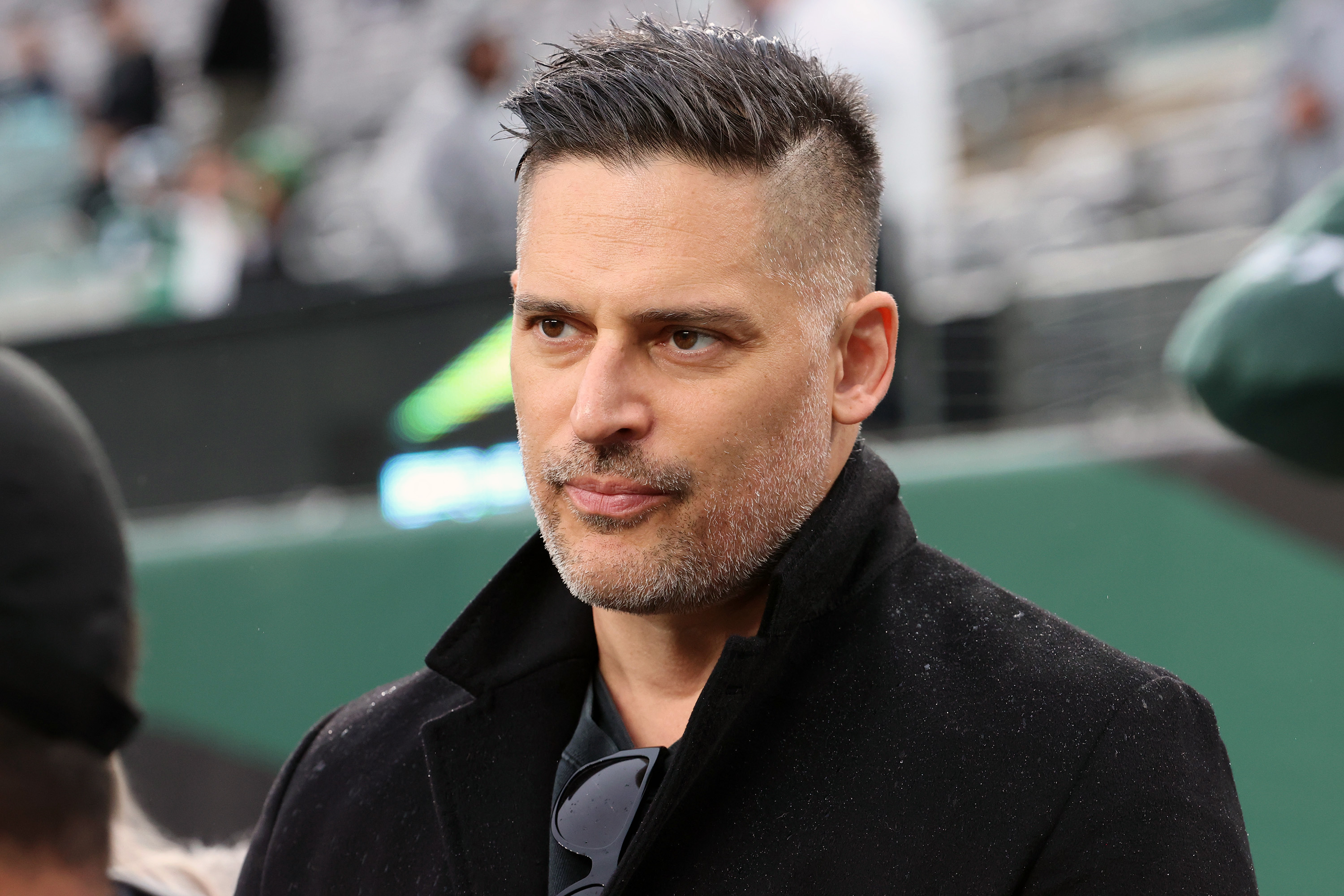 Joe Manganiello stands outdoors in a dark coat, with a serious expression, possibly at an event or gathering